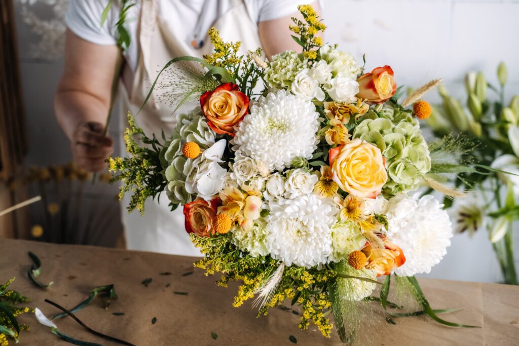 Florist arranging a vibrant bouquet with white chrysanthemums, yellow roses, and mixed seasonal flowers on a wooden table