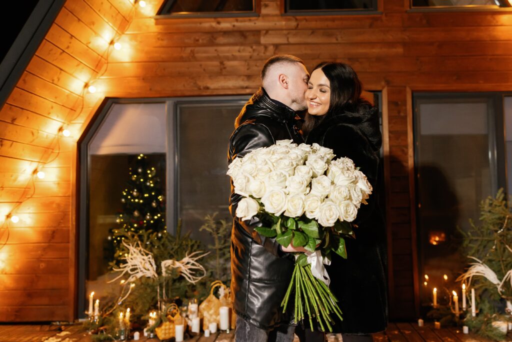 Romantic couple embracing while holding a large bouquet of white roses in a cozy, softly lit setting